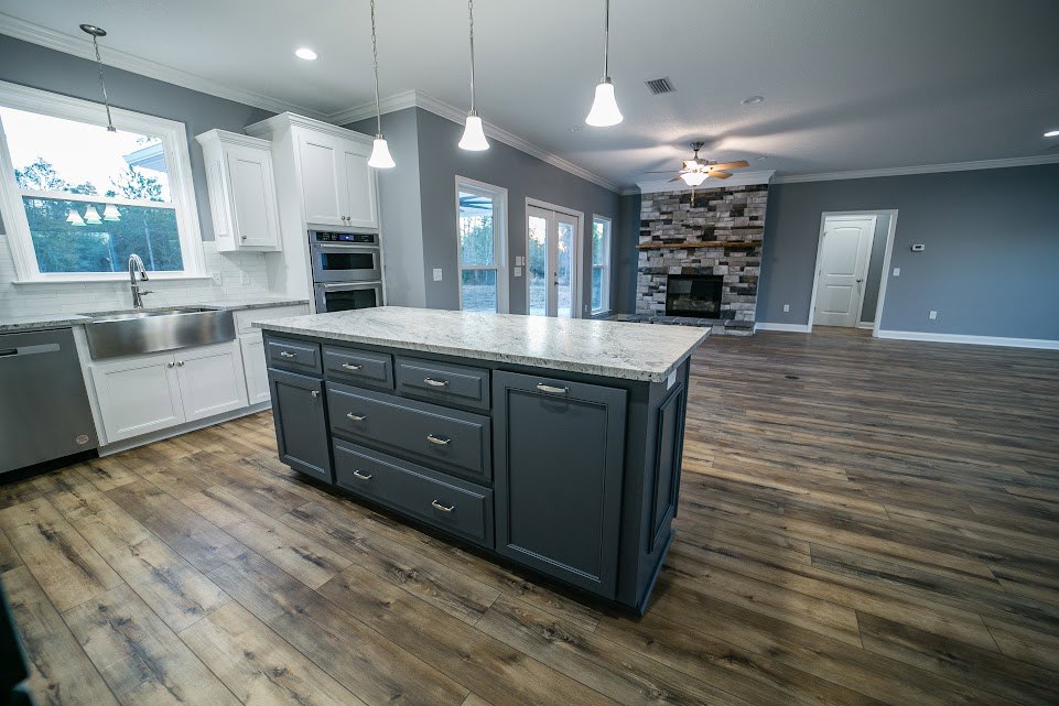 Spacious kitchen featuring a large marble-topped island, white cabinetry with drawers, stainless steel microwave, undermount sink beneath a window, and light wood flooring