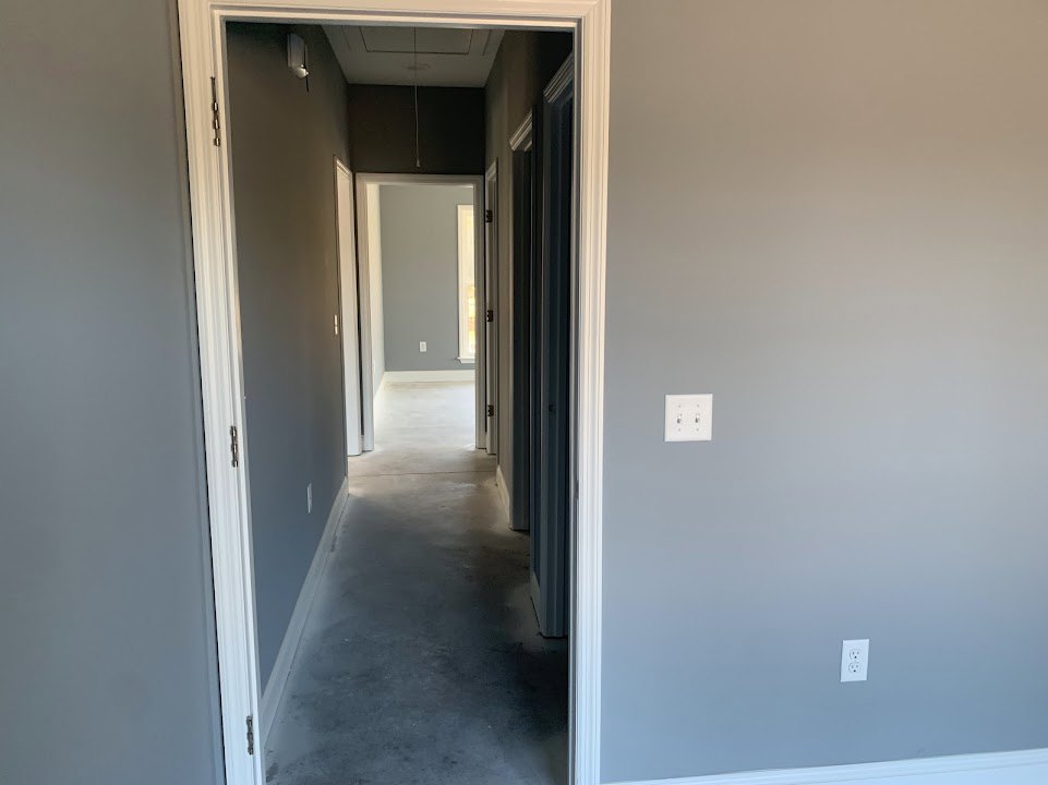 Hallway with gray walls, white trim, white floor, and a white door; light switch on the wall; ceiling light casting illumination on the ground