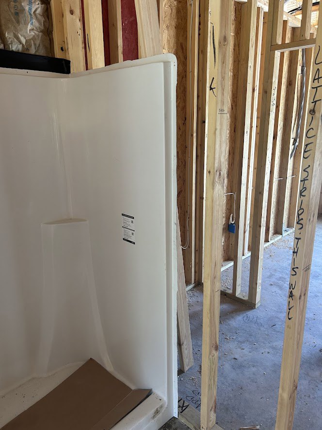 Walk-in shower with white plaster walls, brown rectangular bench, exposed wood framing, and construction materials including a white bag with blue writing.
