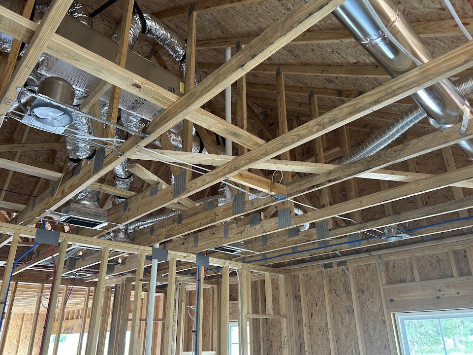 Exposed wood framing with ventilation pipes and clear plastic tubing, white-framed window, metal ductwork, and ceiling beams in a partially constructed home interior