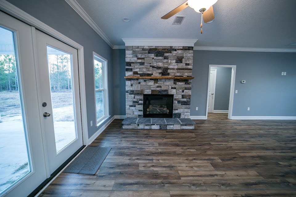 Living room with wood floor, gray rug, white-framed door, stone fireplace topped by a wood mantel, and ceiling fan.