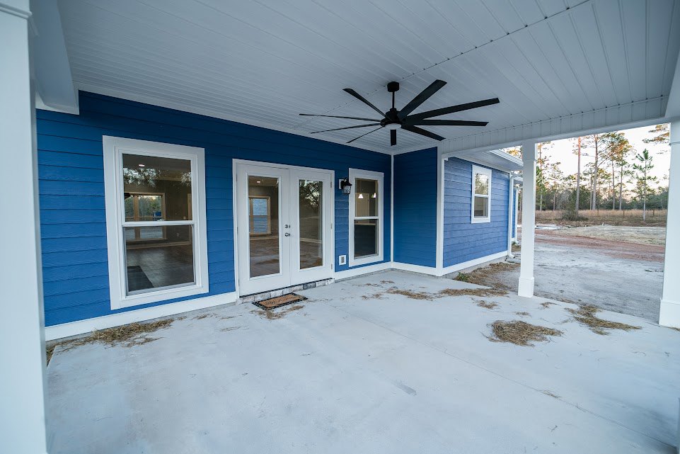 Blue siding with white trim, double glass doors, ceiling fan on covered porch, concrete floor, windows illuminated from inside, snow on ground.