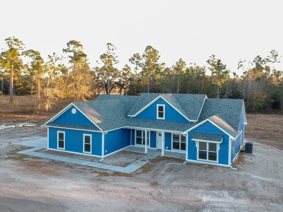 Blue house with white trim and windows, white front door, concrete driveway, mature trees in the background, clear sky above.