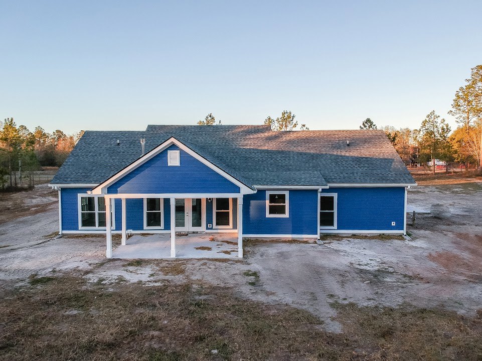 Blue siding house with white trim and pillars, front porch, illuminated window, gabled roof, mature trees in background.