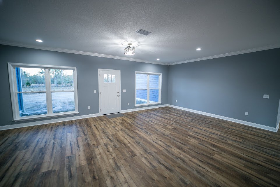 Bright room with wide wood flooring, white walls, large windows showing blue sky and green yard, white door with glass panel.