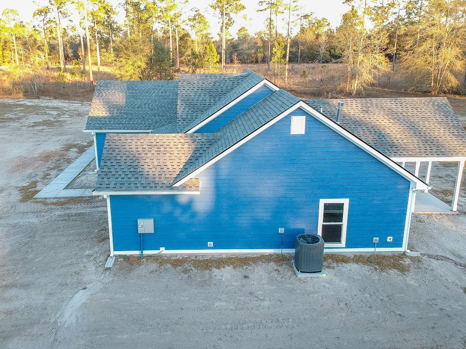 Blue siding house with white roof, large concrete driveway, white-framed windows, grey air conditioner unit, trees in background