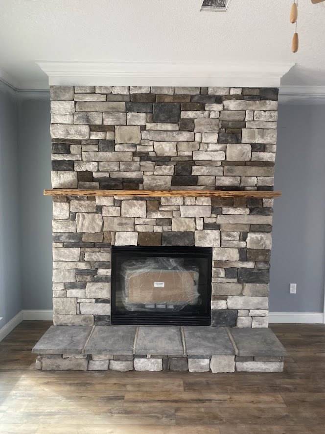 Stone fireplace with wood mantel set against a brick accent wall, hardwood flooring, fire screen, and wood burning stove in a modern living room.
