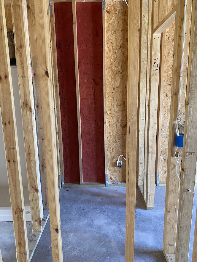 Wood framing and beams on a concrete floor, red wall with white trim, exposed blue electrical box and white wires, unfinished interior construction.
