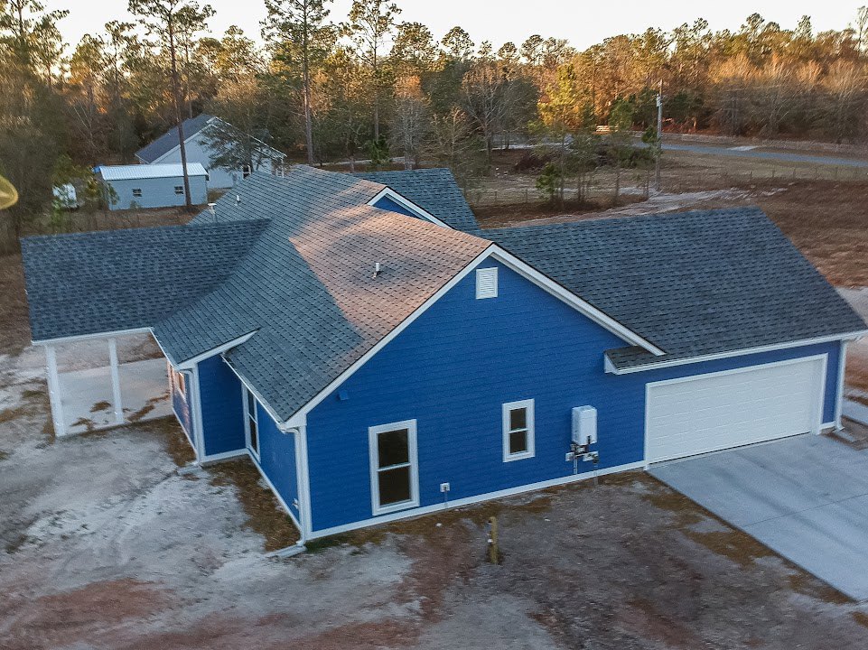 Blue house with white roof and trim, attached garage, white-framed windows, person standing beside garage door, tree and sky in background.