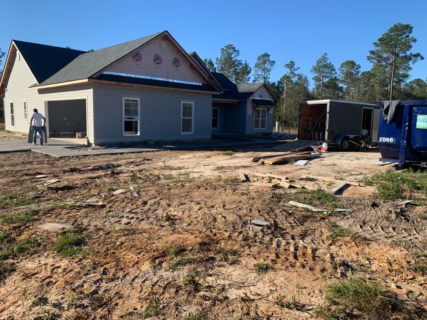 Partially built house with exposed framing and roof, construction trailer parked on dirt lot, scattered wood pieces, man standing near unfinished garage door, trees and blue sky in