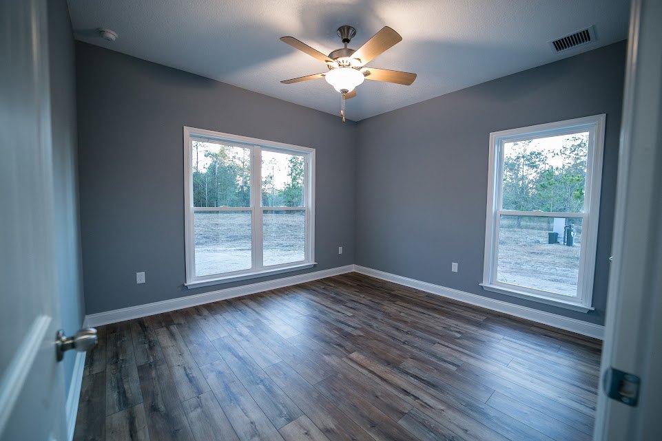 Ceiling fan with light fixture, multiple windows overlooking trees, wood flooring, white walls, partial view of faucet