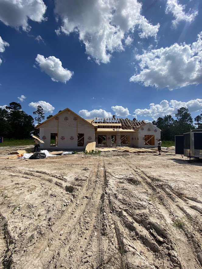 Wood-framed house under construction on dirt lot, ladder leaning against exterior, man standing nearby, tire tracks visible in foreground, blue sky overhead
