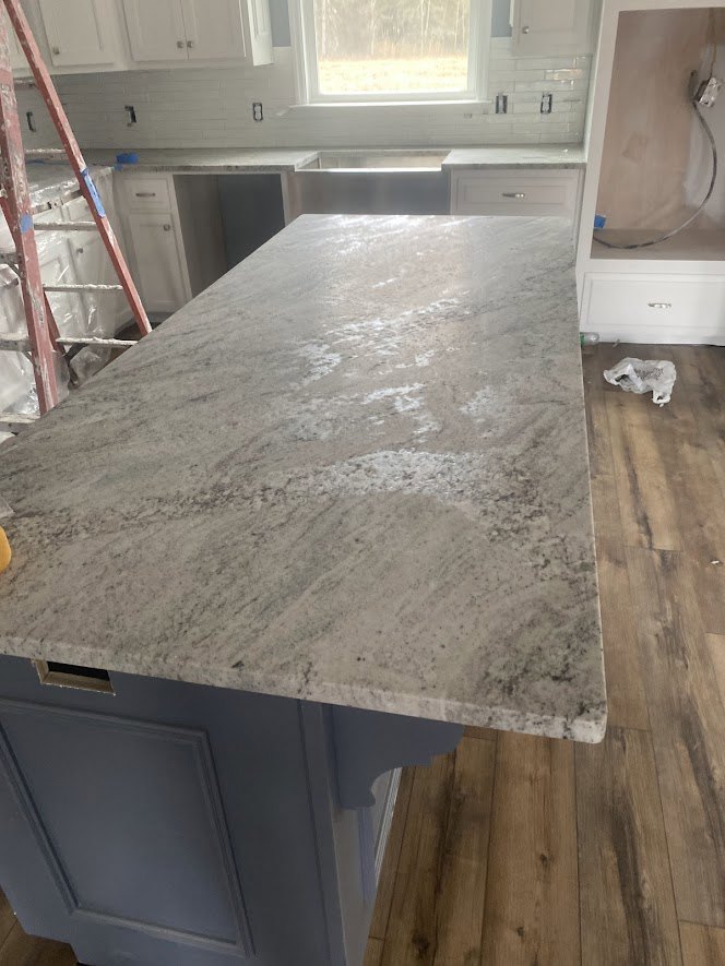 Kitchen under renovation with plywood countertop, white plastic bag, wire along plaster wall, paint-splattered ladder, wood flooring, and window overlooking field and trees