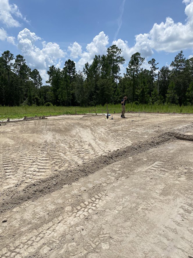 Man standing on a dirt lot surrounded by trees, blue sky overhead, tire tracks visible in soil