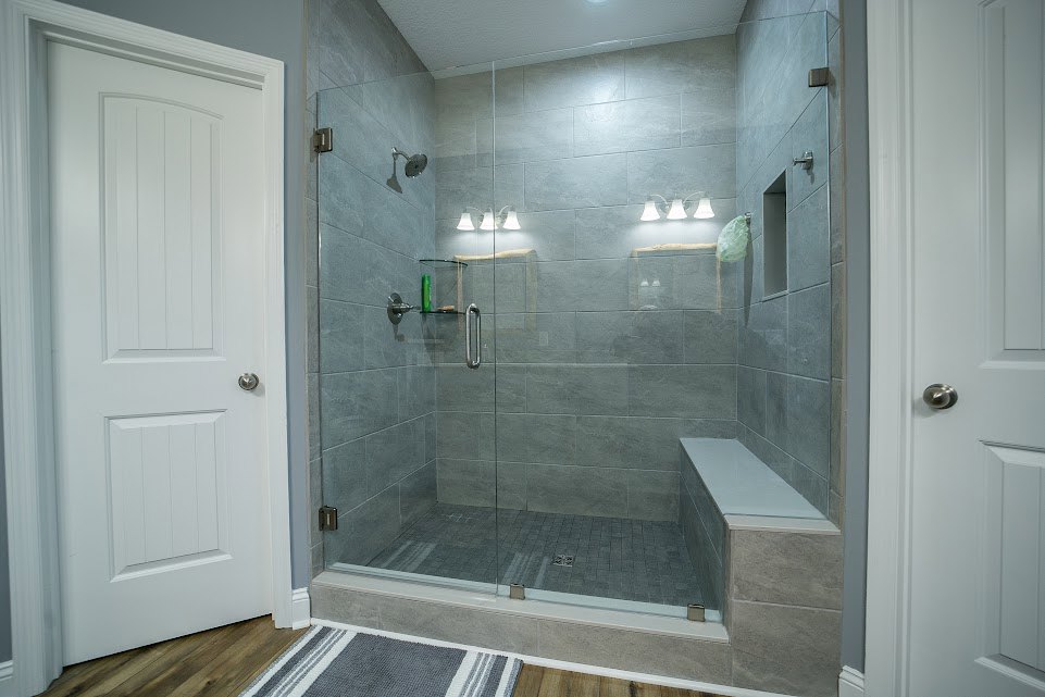 Glass shower enclosure with built-in bench, light tile walls and floor, white door with silver knob, textured rug in foreground