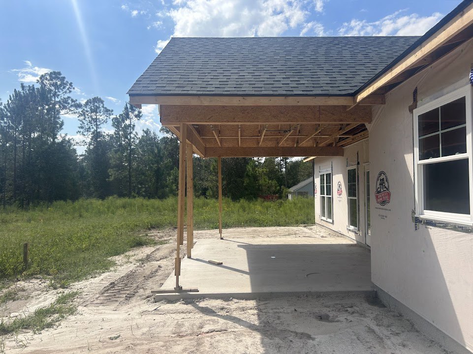 Partially built house with exposed framing, white window frame, unfinished roof, concrete porch, grassy yard, and mature trees in the background
