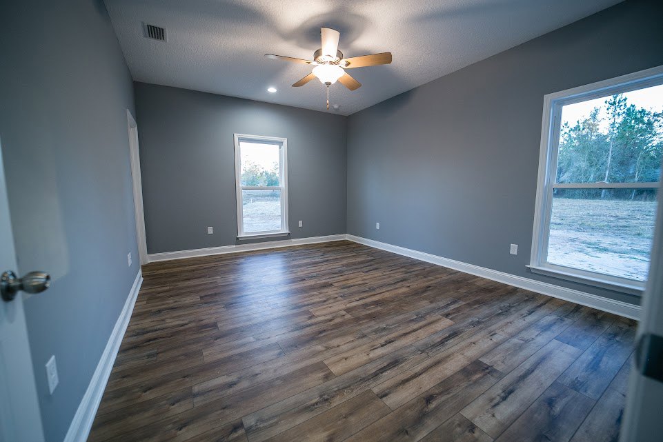 Wood flooring in a bright room with a ceiling fan and light fixture, white walls, large window showing green trees outside, close-up of a brushed metal door knob