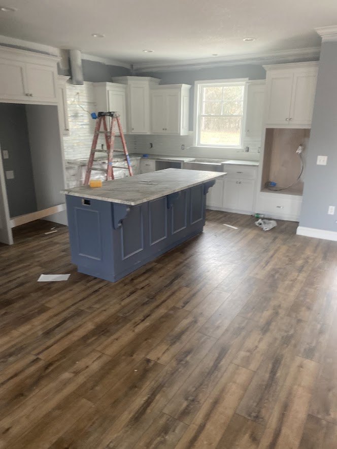 Kitchen with wood flooring, marble-topped island, white cabinetry, window with white frame and curtain, and a ladder with blue roller leaning against a white wall