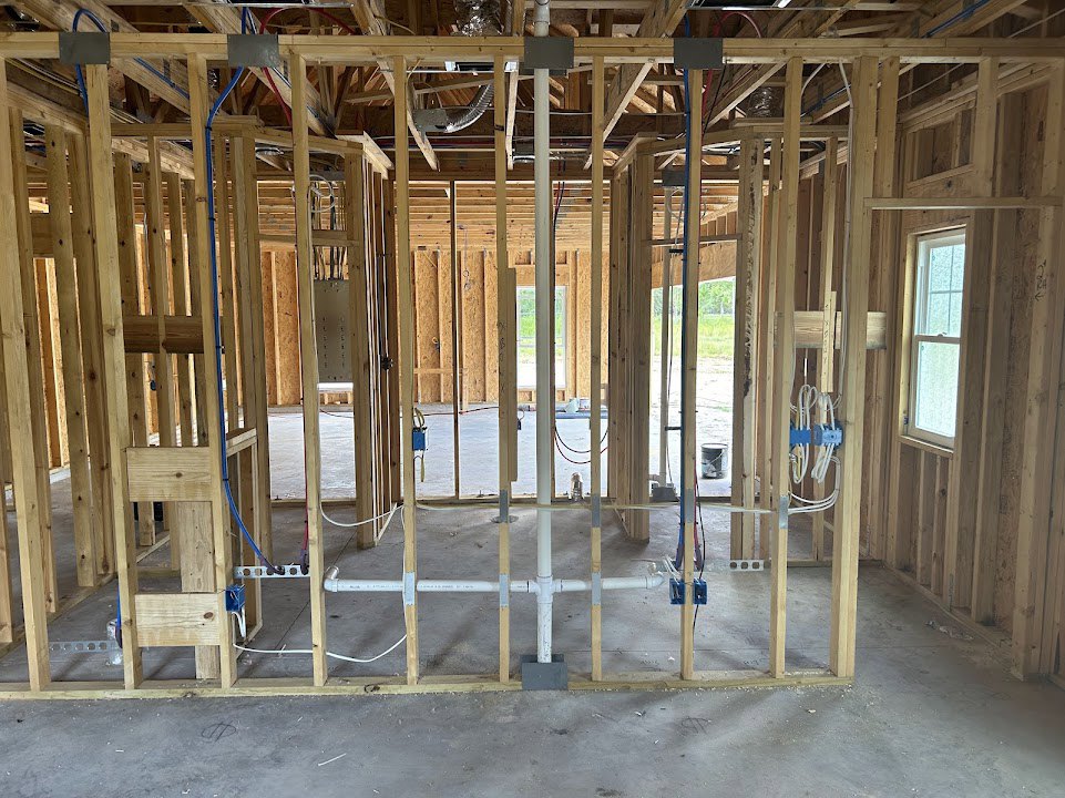 Exposed wood framing with white-framed window, blue and white electrical wires, white pipe along studs, concrete floor, and metal support pole