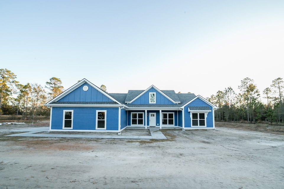 Blue house with white trim and blue roof, white-framed windows, dirt road in front, surrounded by trees and forest