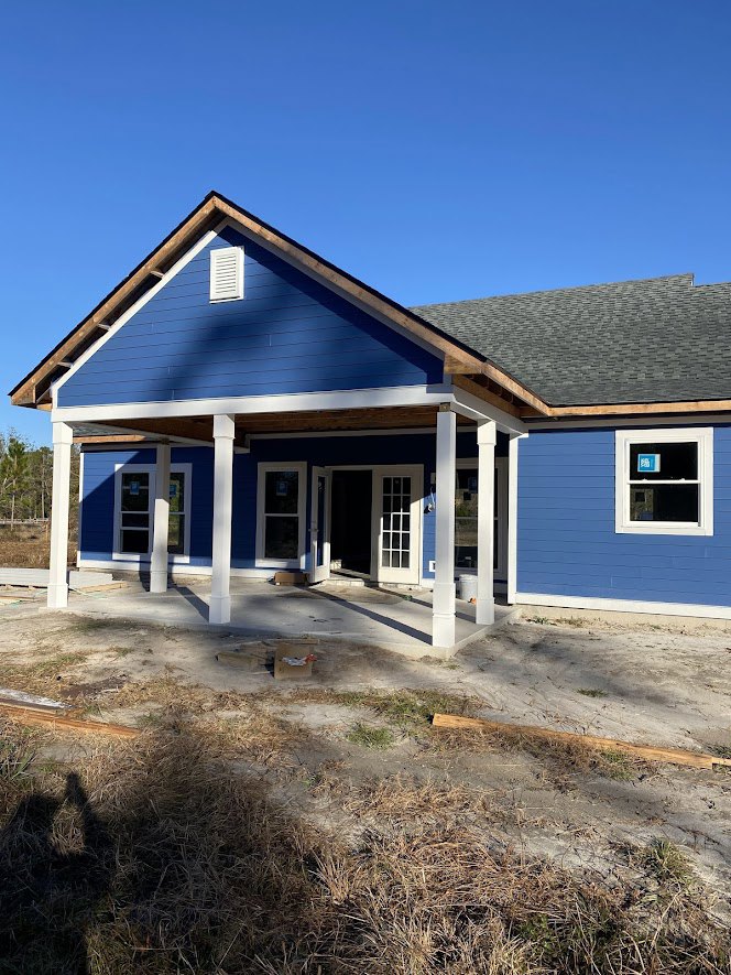 Blue siding house under construction with white pillars on porch, windows featuring blue signs, and unfinished landscaping with grass in foreground.