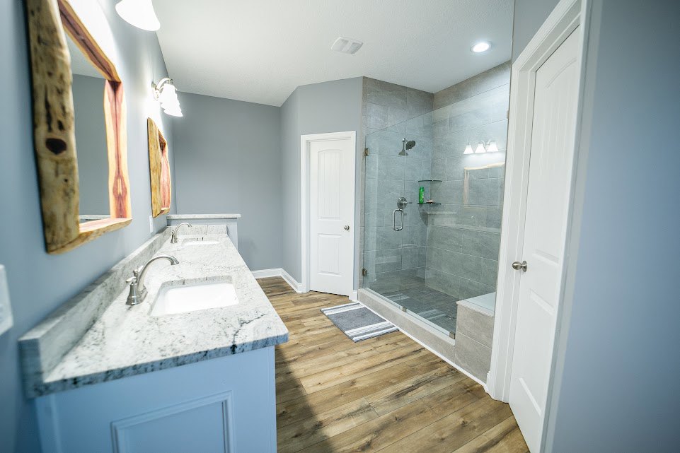 Bathroom with frameless glass shower enclosure, double white sinks set in a light countertop, large wall mirror framed in wood, wood flooring, and neutral tile accents.