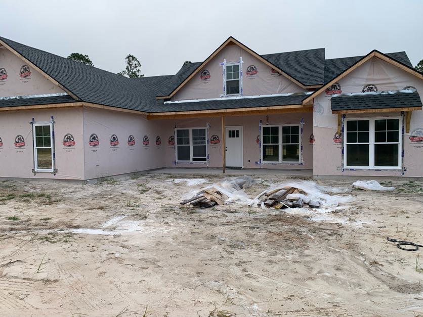 Partially built house with white-framed window, taped exterior walls, exposed roof, and piles of wood and plastic on dirt yard