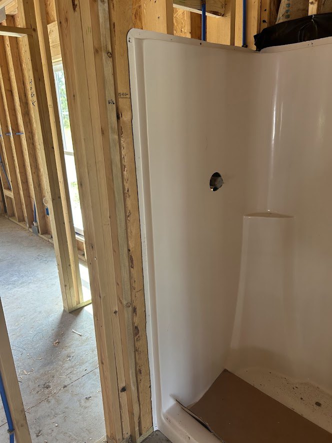 White tile shower with built-in niche, black shower fixture, brown wood box in bathtub, unfinished plywood frame on floor, plastered walls.