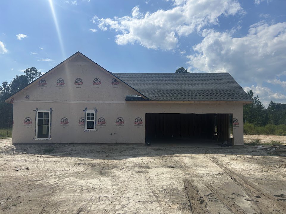 Partially built house with white-framed windows, exposed roof trusses, attached garage, dirt lot with tire tracks, blue sky and scattered clouds
