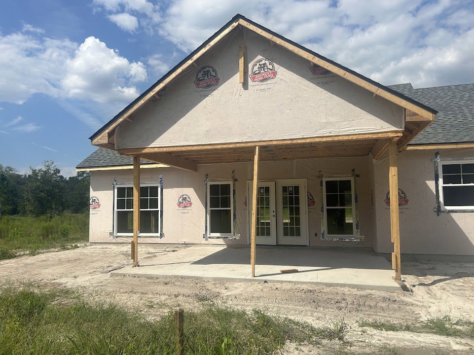 Partially built house with exposed wooden framing, fenced yard with green grass, white-framed windows, wooden pole near window, person visible inside, logo displayed on exterior