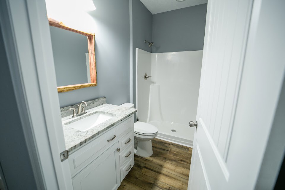 White toilet and rectangular sink with silver faucet, wood floor, white cabinetry, and partial view of door frame in modern bathroom.