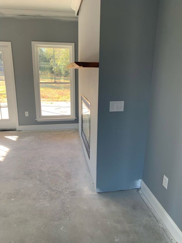Living room with concrete flooring, plaster walls, built-in fireplace, large window with outdoor view, white door frame, and sunlight streaming onto a white surface.