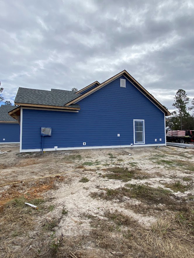 Blue house with horizontal siding, gabled roof, and white trim, set beside a dirt field and surrounded by leafy trees under a cloudy sky.