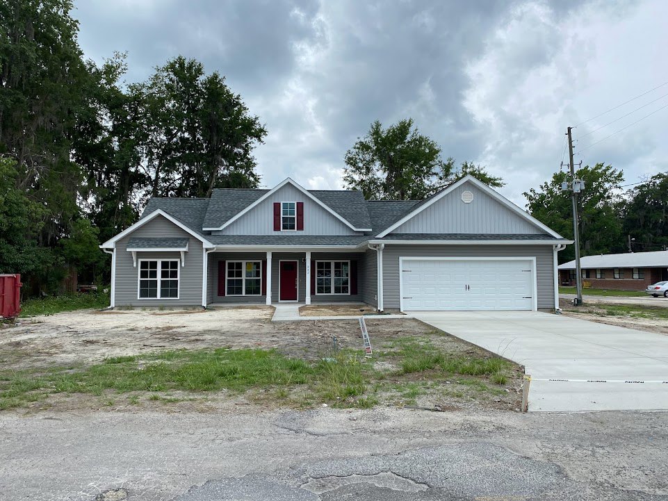 Two-story house with red front door, white garage door with black handles, driveway bordered by grass and fence, group of mature trees, white-framed windows, cloudy sky overhead