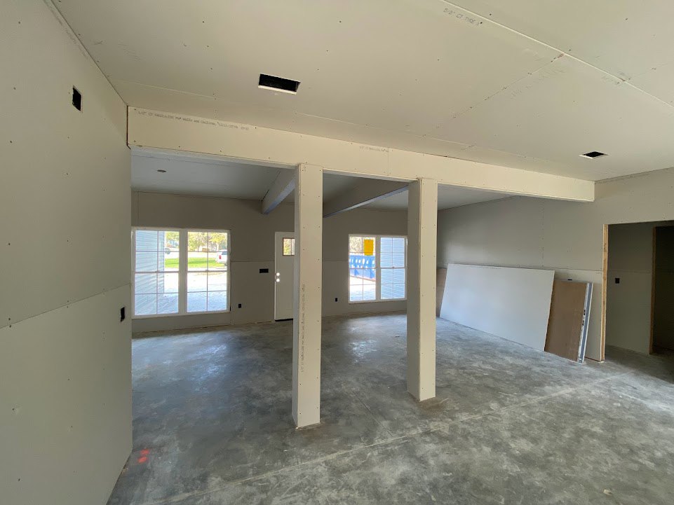 Spacious room featuring concrete floor, white plaster walls with wooden framing, tall pillars, large windows overlooking a green yard, and a close-up of a wooden door; person