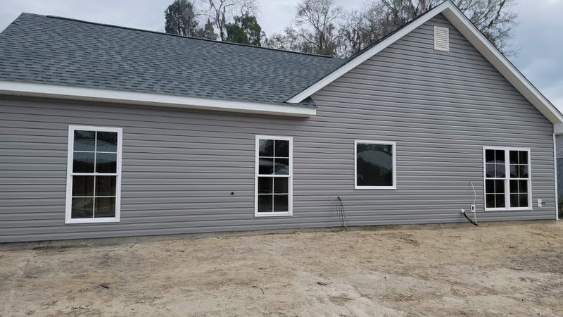 Grey house with white trim and multi-pane windows, dirt driveway in front, metal siding visible on lower exterior.