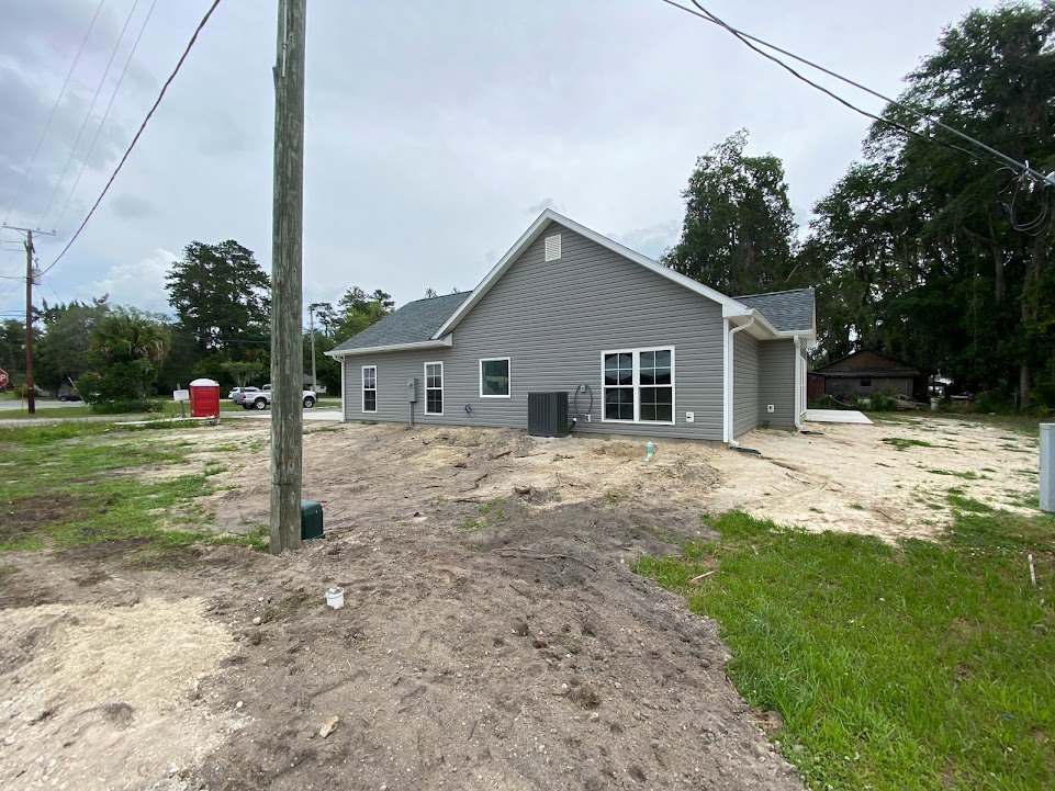 Two-story house with grey roof, multi-pane windows, and a dirt patch in the front yard, surrounded by grass, trees, and power lines under a partly cloudy sky