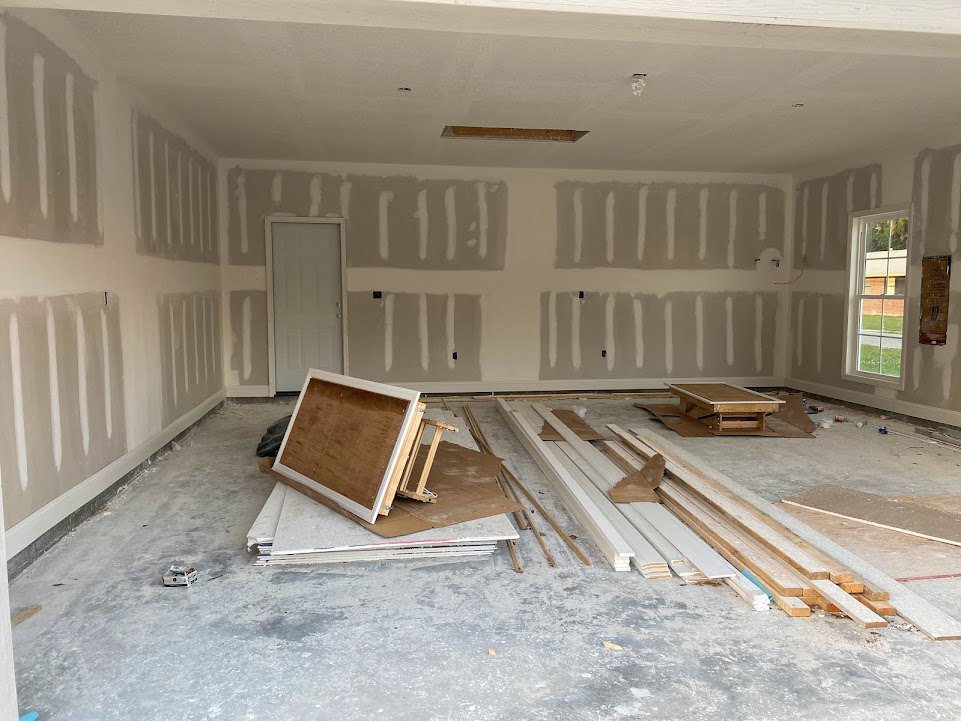 White door with wooden frame leaning against a plaster wall, scattered wood beams on cardboard-covered floor, window reveals brick building outside, table with white edge in