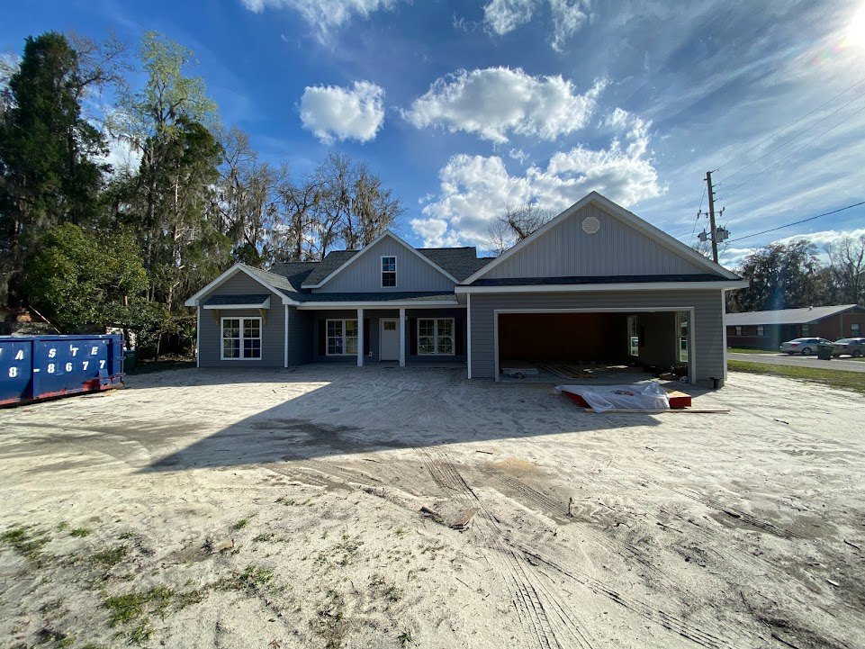 Two-story house with attached garage, white-framed windows, blue shipping container with white numbers beside driveway, tire tracks in sandy ground, trees and partly cloudy blue