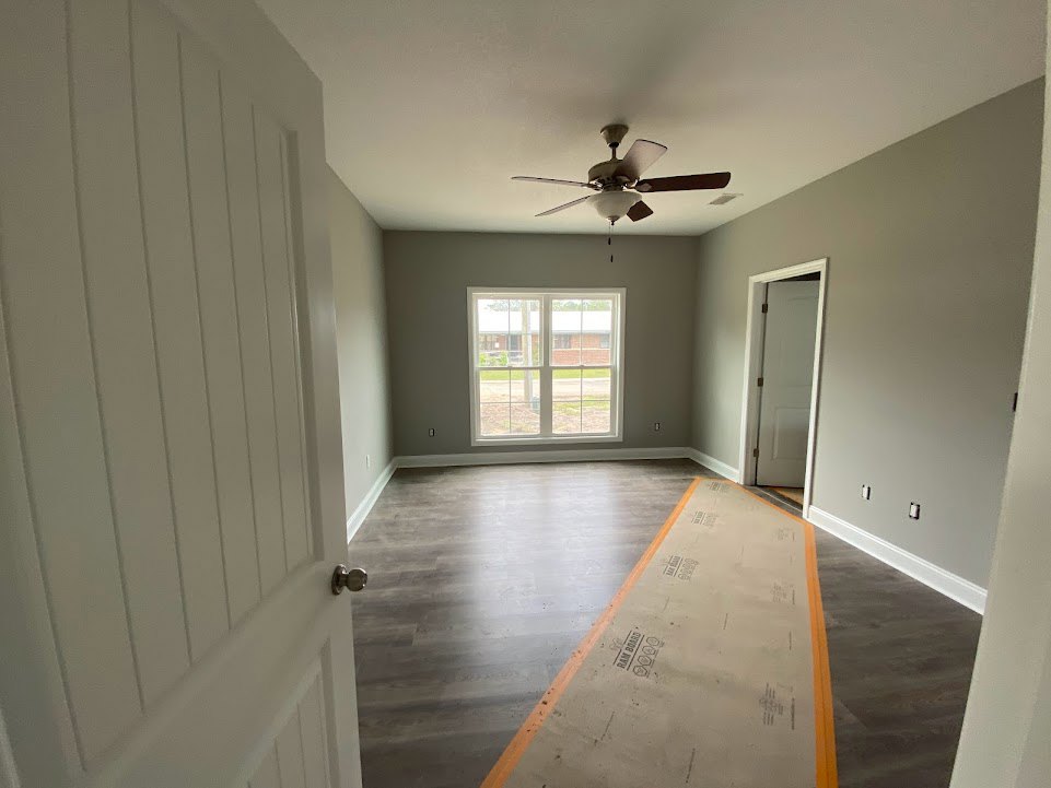 Carpeted room with a ceiling fan and light, white paneled door with metal hinge, window overlooking yard, neutral walls and grey-orange flooring