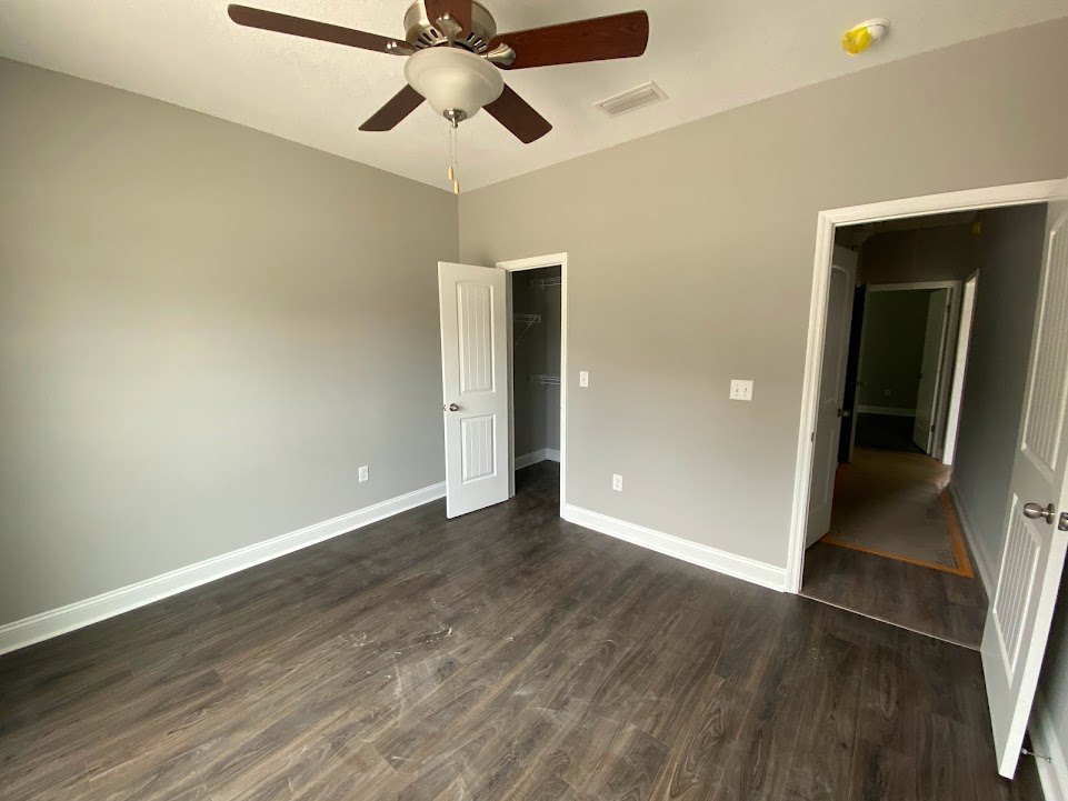 Ceiling fan with light fixture above wood flooring, white baseboards, and white doors with silver knobs; hallway visible with two doors and neutral plaster walls.