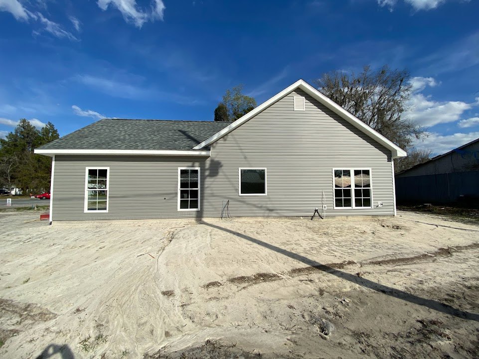Tan siding house with white-framed window, dark roof, and dirt yard under blue sky