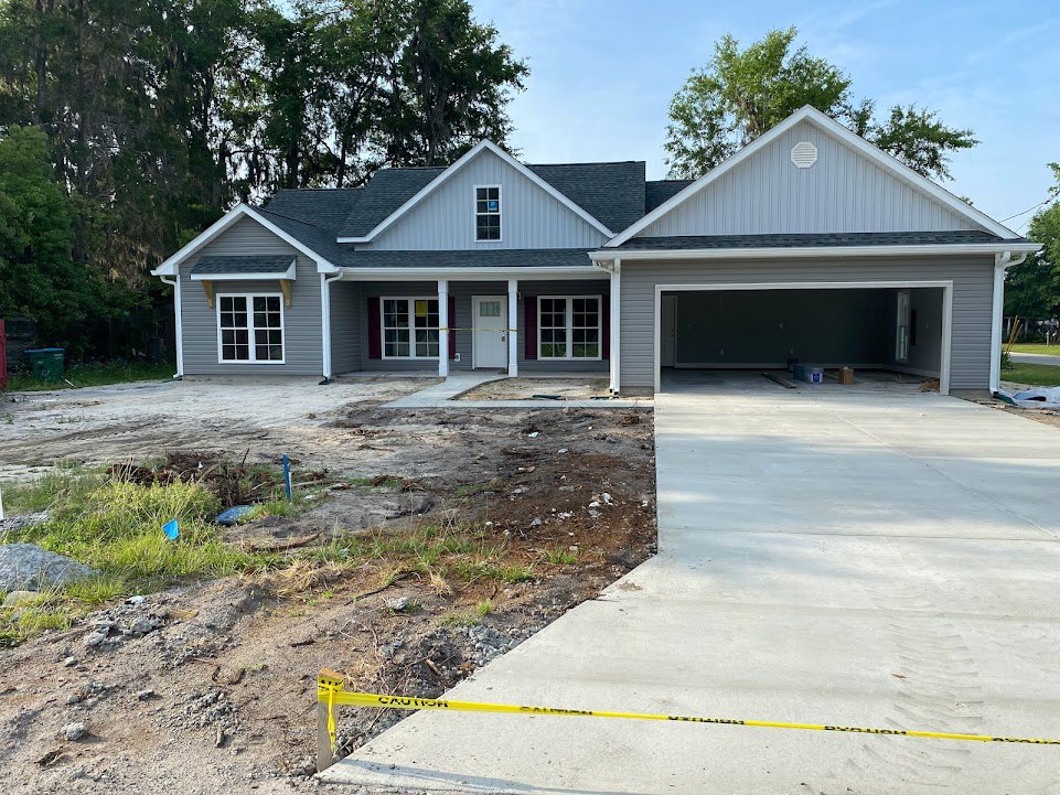 Two-story house under construction with exposed framing, white window frames, attached garage, concrete driveway, and yellow caution tape along the sidewalk.