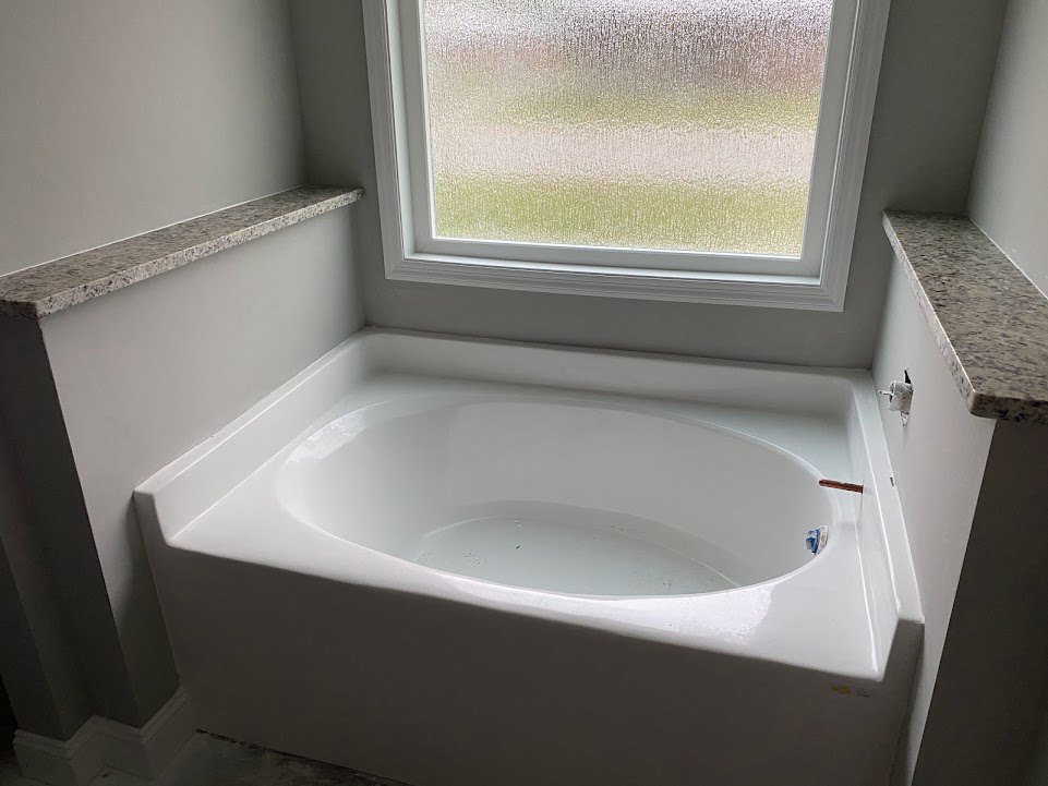 White freestanding bathtub filled with water, set on light tile floor beside frosted glass window; nearby countertop with chrome faucet and bathroom accessories visible.