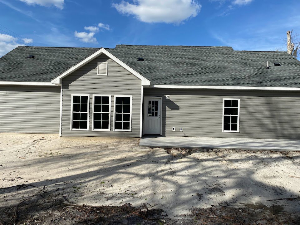 Two-story home with gray siding, white trim, row of windows, white front door with glass panel, paved driveway, gabled roof, and cloudy sky overhead