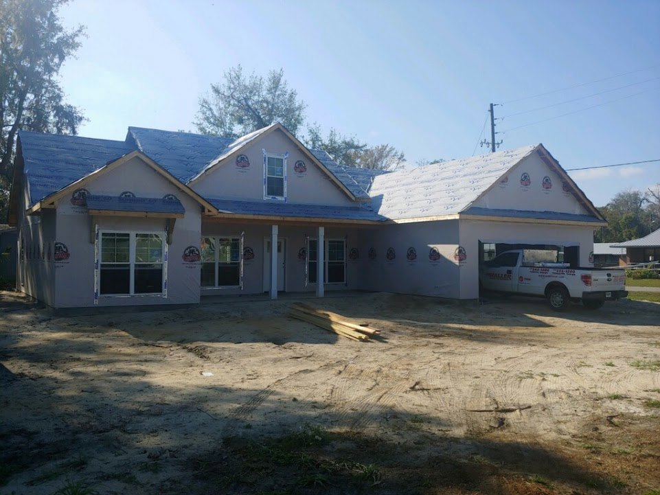 Partially built house with exposed framing, white truck parked in front, white-framed window, pile of lumber on dirt, shingled roof, trees and sky in background