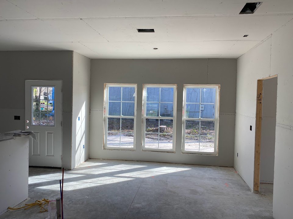 Spacious room featuring large windows with white frames, concrete flooring, white plaster walls, and a white door with a wooden frame overlooking a landscaped yard.