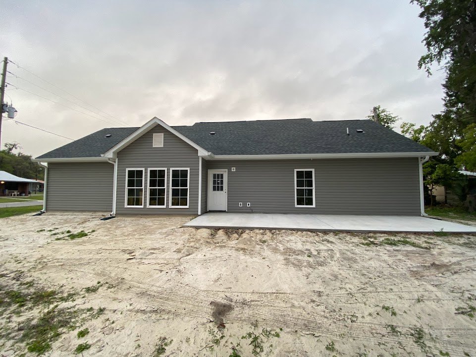 Grey two-story house with white trim, row of windows, covered porch, dirt driveway with tire tracks, surrounded by trees under cloudy sky