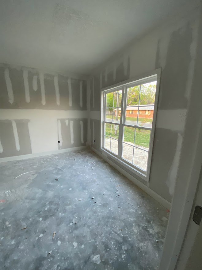 Sunlit room with smooth plaster walls, large window framed in white, subtle shadows cast across grey surfaces, and exterior building visible through glass.