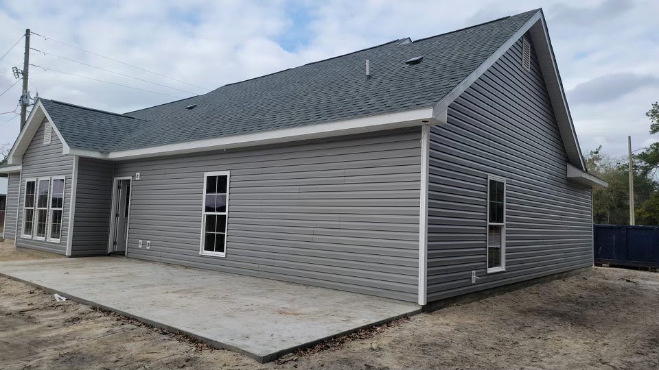 Grey siding exterior with white-framed window, concrete patio in foreground, blue recycling bin near curb, partly cloudy sky overhead